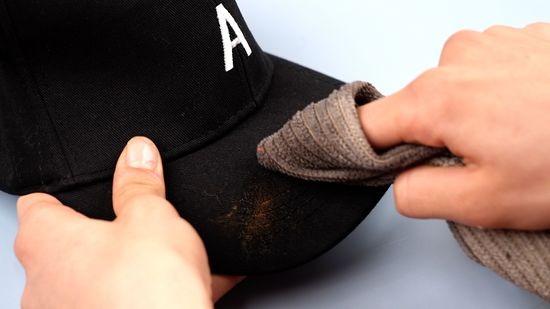 Manos limpiando una gorra de béisbol negra con un trapo, quitando la suciedad visible del ala.