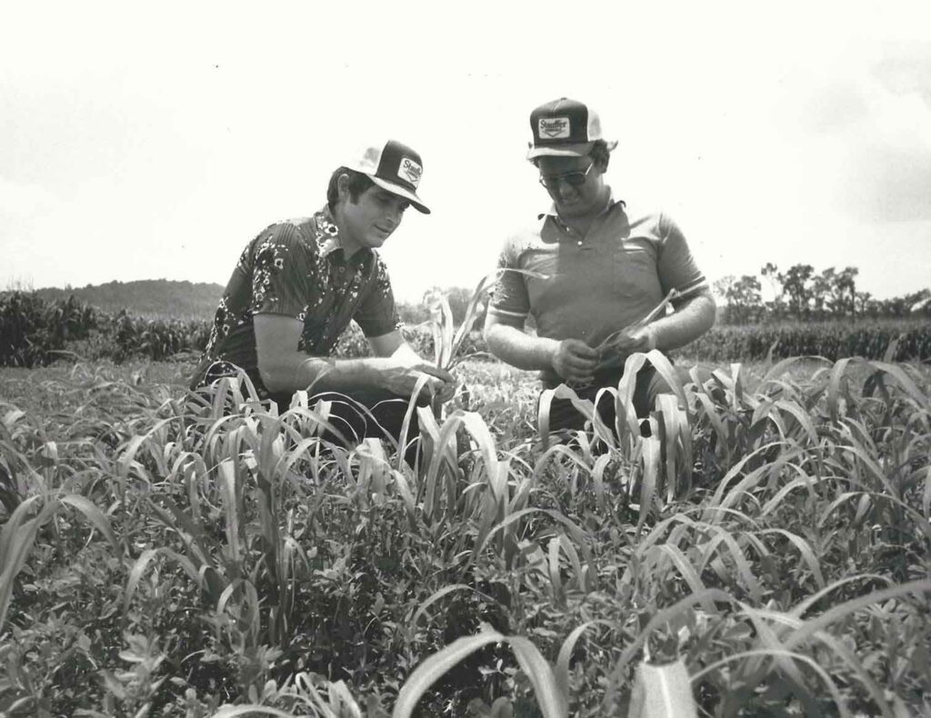 Dos hombres con sombreros de camionero examinan los cultivos en un campo, captados en una foto en blanco y negro de una década pasada.