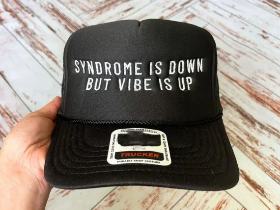 Gorra negra de camionero con texto bordado en blanco en el que se lee "Syndrome is down but vibe is up" (El síndrome ha bajado pero las vibraciones han subido), sostenido por una mano sobre fondo de suelo de madera.