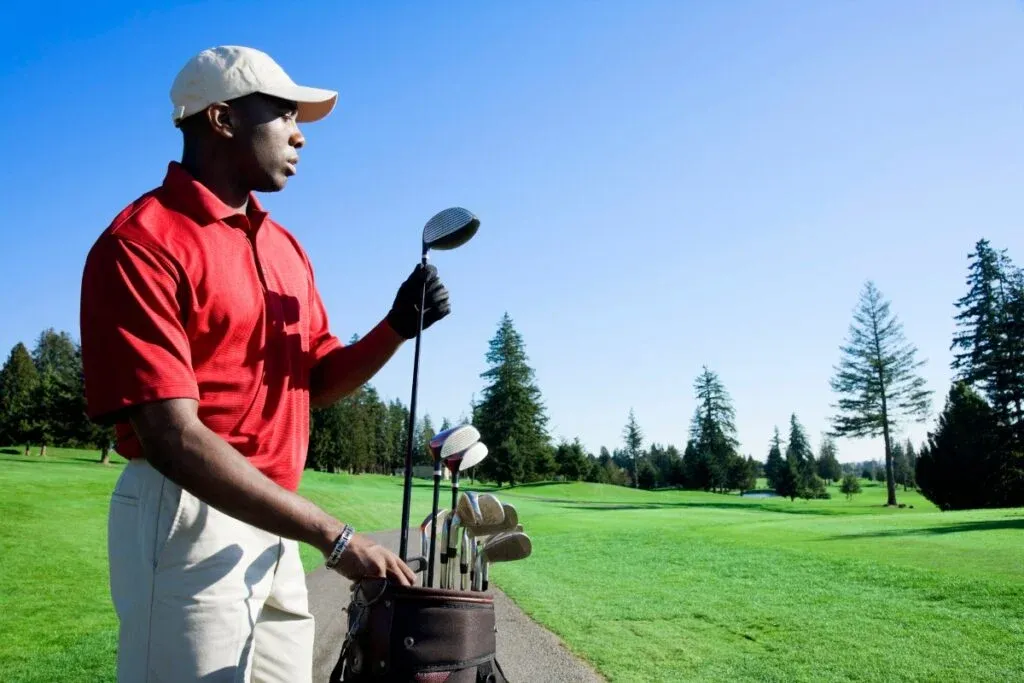 En golfare i röd polo och vit keps står på en bana och väljer en klubba ur sin golfbag under klarblå himmel.
