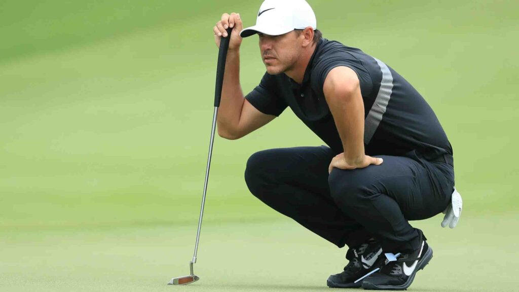 Male golfer in black attire and white cap squats on the green, studying the line before a putt with intense concentration.