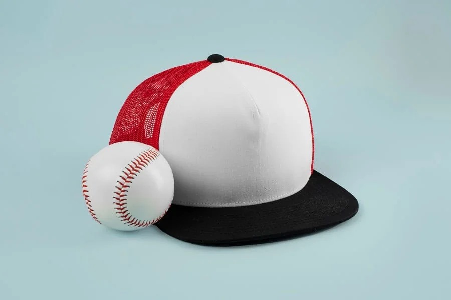 White baseball cap with red mesh and black brim next to a baseball ball.