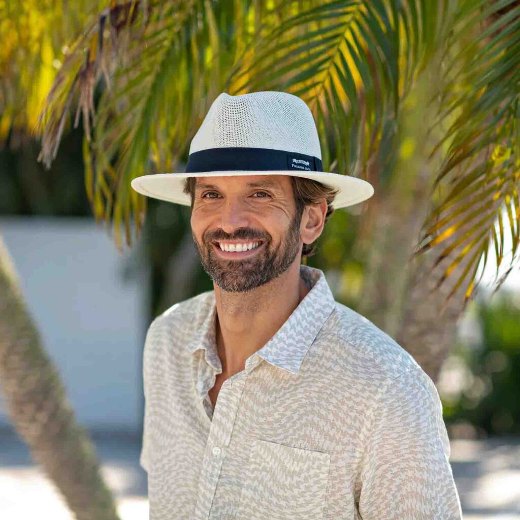 Smiling man wearing a white Panama hat and patterned shirt under palm trees.