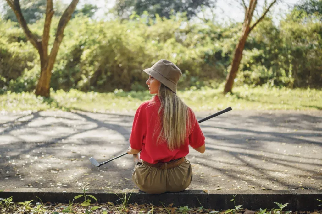 Female golfer in a red shirt and bucket hat sits on a curb holding a golf club, surrounded by trees in a sunlit park.
