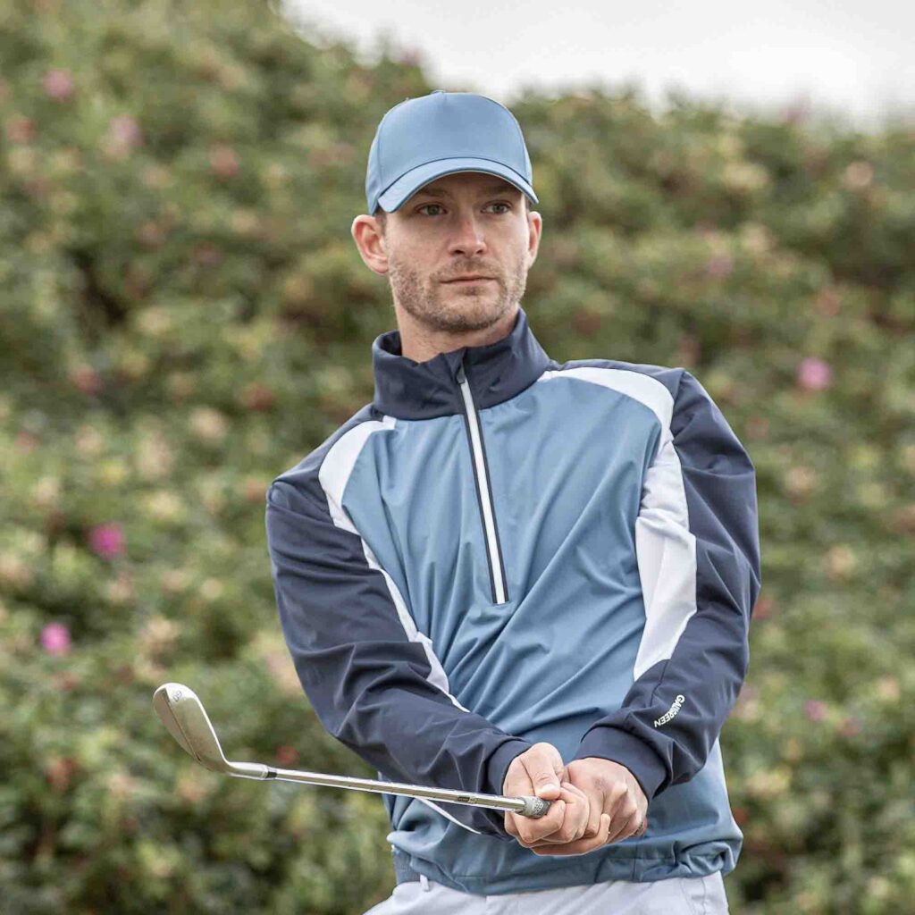 Man wearing light blue golf cap and windbreaker jacket while swinging club outdoors