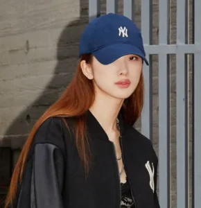 Young woman in blue baseball cap and black jacket posing outdoors.