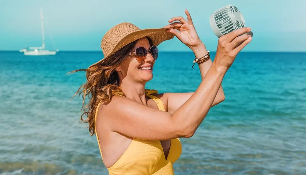 Smiling woman wearing straw hat by the sea holding a handheld fan.