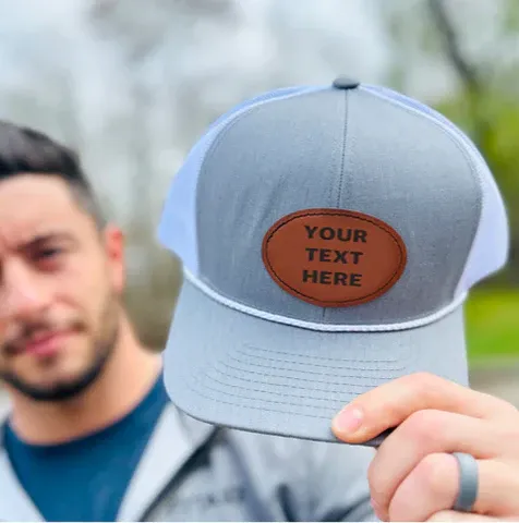 En man håller i en grå och vit truckerkeps med en brun läderlapp där det står "YOUR TEXT HERE", perfekt för kundanpassning.