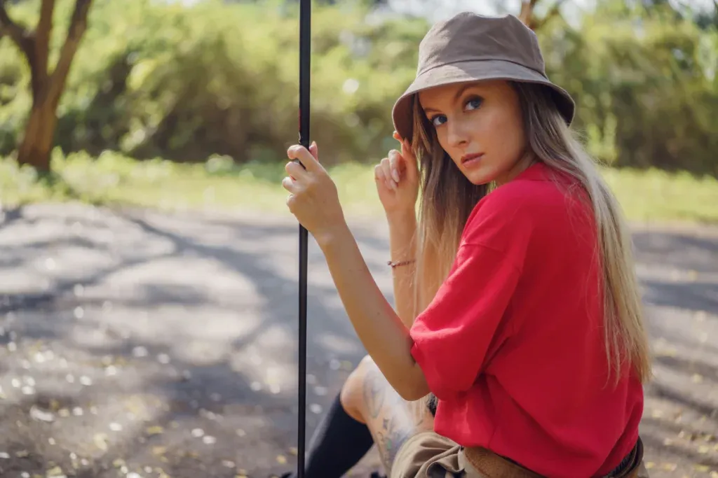 Young woman in a red shirt and bucket hat sitting with a golf club, gazing at the camera in a shaded outdoor setting.