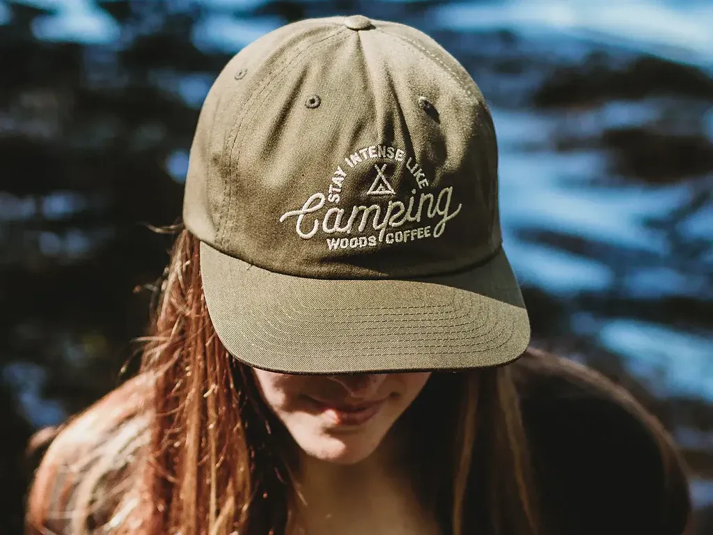 Woman wearing olive green cap with “Camping Woods Coffee” embroidered logo.