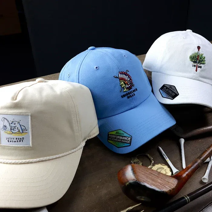 Tre snygga broderade golfhattar - creme, blå och vit - utställda på en träyta bredvid vintage golfklubbor och tees.