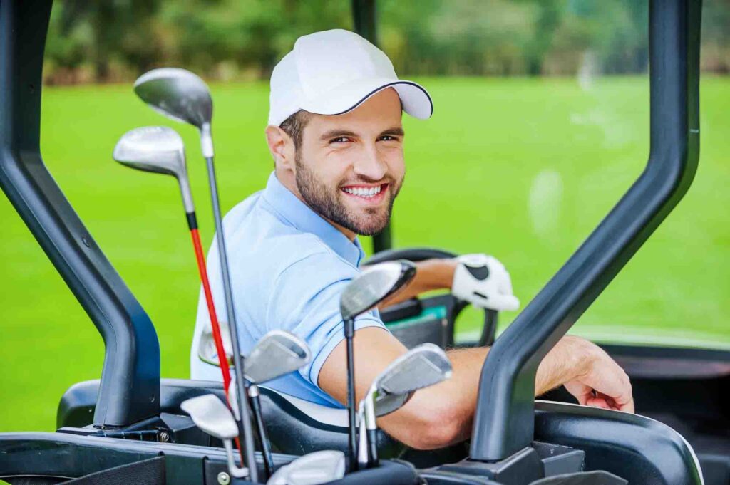 Smiling man in white cap driving golf cart with clubs on a green course