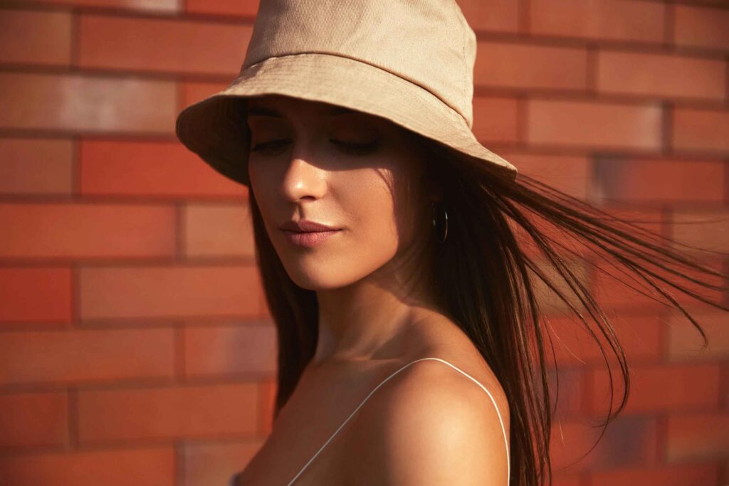 Vrouw met lang donker haar draagt een beige emmerhoed en hoepeloorbellen tegen een rode bakstenen muur.