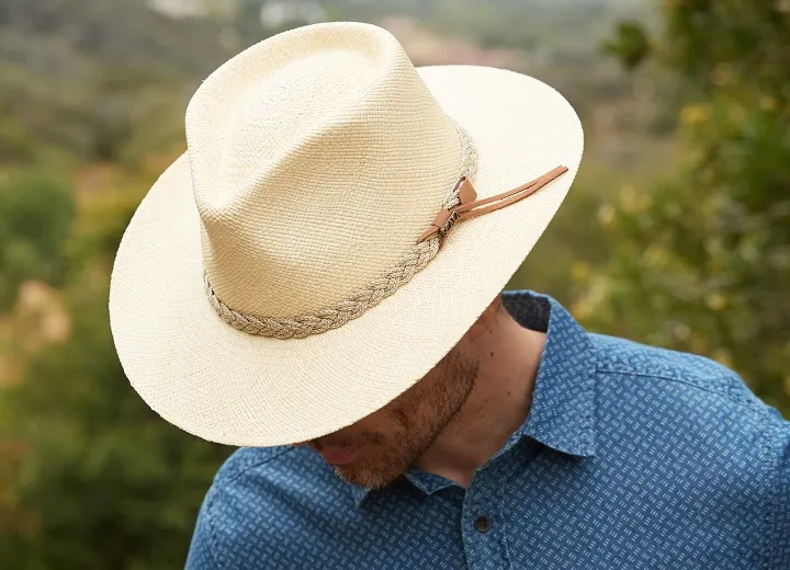 Man in blue shirt wearing cream straw hat with braided band outdoors.