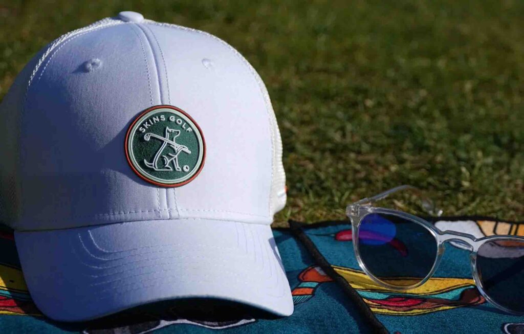 White Skins Golf cap with green logo patch placed on grass beside sunglasses