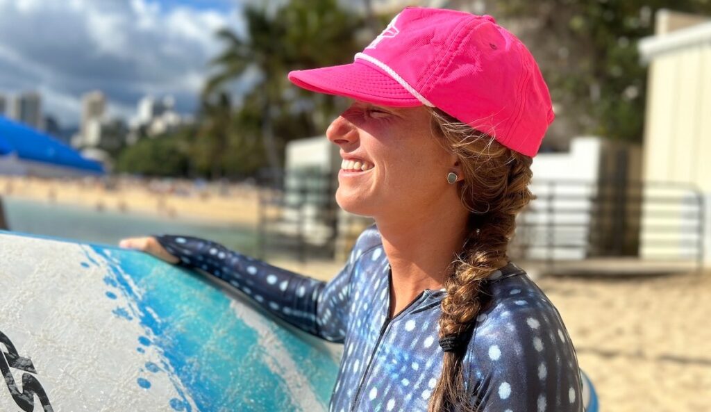 Smiling woman in pink cap holding surfboard on sunny beach with city background.