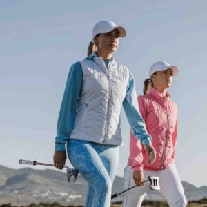 Två kvinnor i snygga golfkläder och vita kepsar går självsäkert med putters på en golfbana.