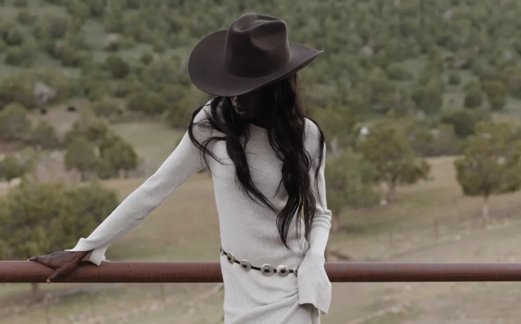 Woman wearing dark wide brim hat and white dress, leaning on fence in outdoor setting