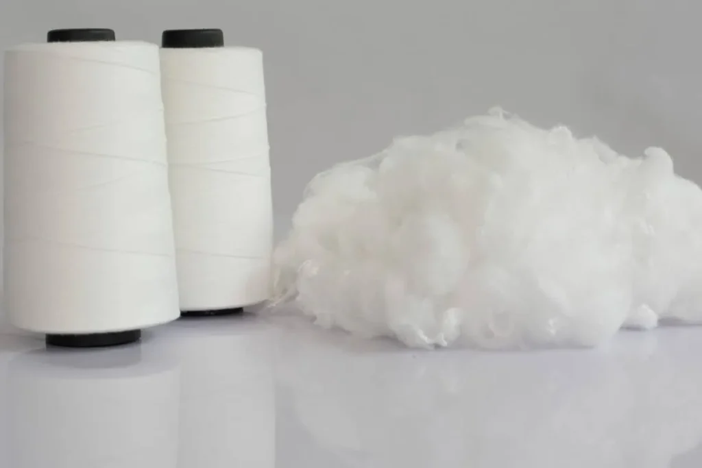 Two white spools of polyester thread next to fluffy polyester fiber on reflective surface.