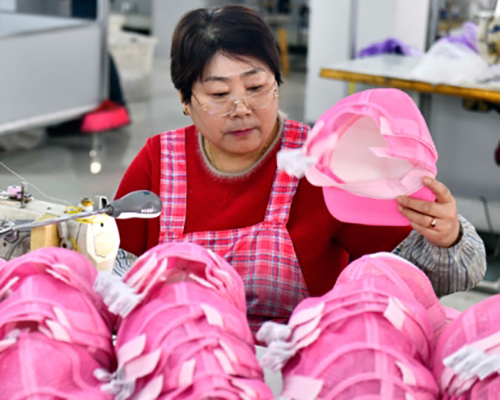 Woman sewing pink caps in factory with piles of caps nearby.