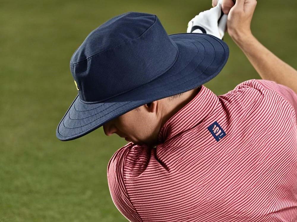 Golfer wearing navy wide brim hat and red striped shirt swinging on green field.