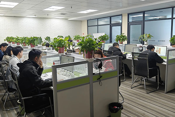 Employees working at desks with computers and plants in office cubicles.