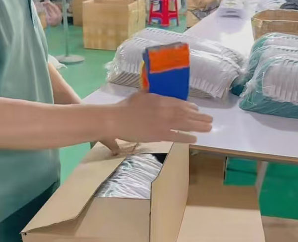Worker packing multiple baseball caps into cardboard boxes on table.