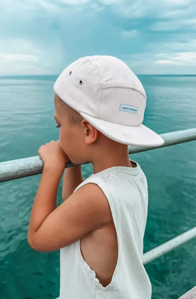 Child wearing a white five-panel cap backwards, looking over railing at blue ocean