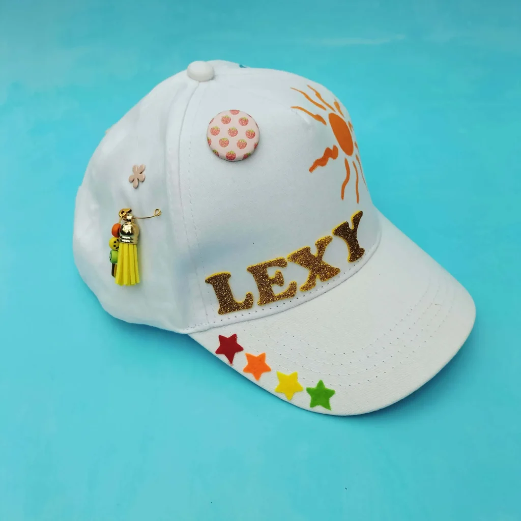 White cap decorated with name, sun, stars, and tassel charm on blue background.