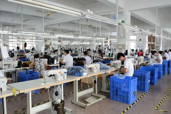 Workers sewing clothes at machines in a large factory with bright lighting.