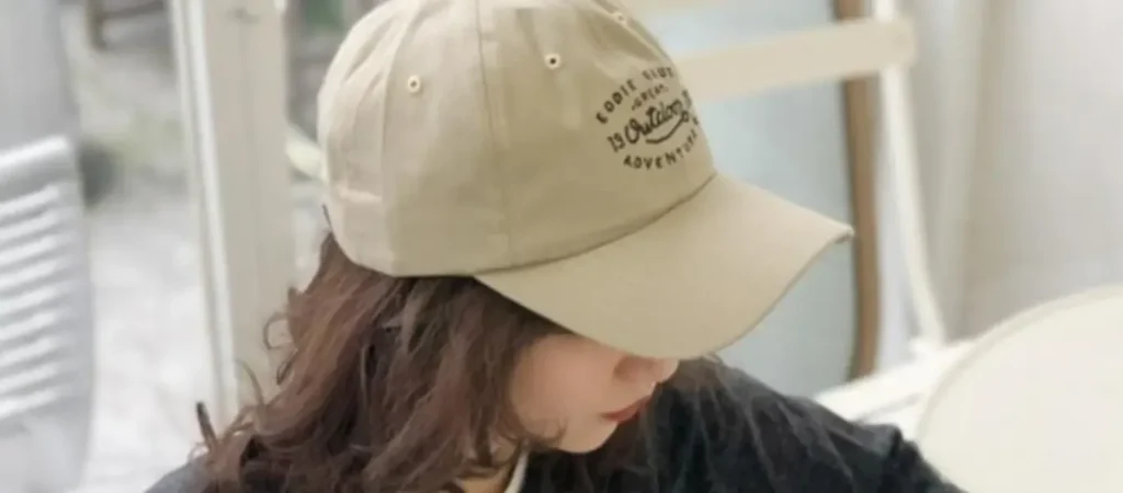 Woman wearing beige baseball cap with embroidered text sitting indoors.
