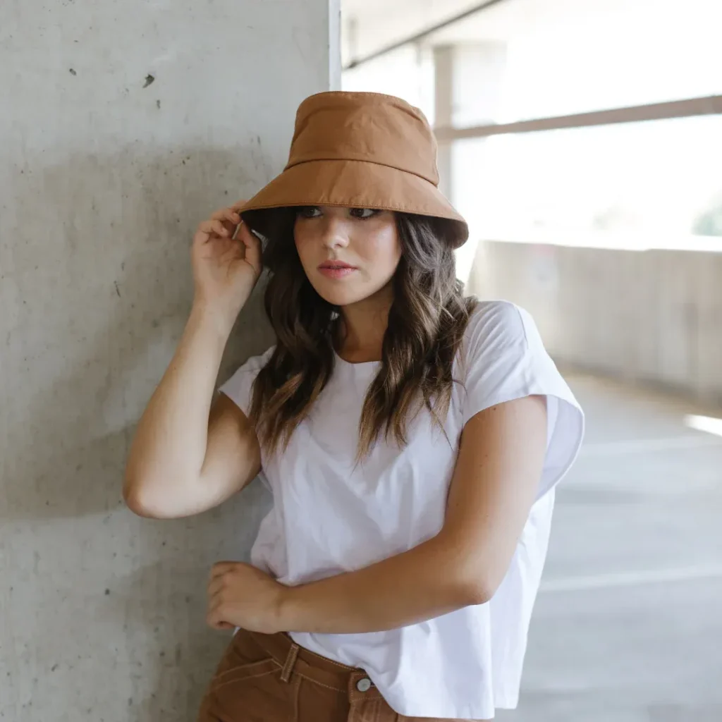 Woman adjusting brown bucket hat leaning against concrete wall.