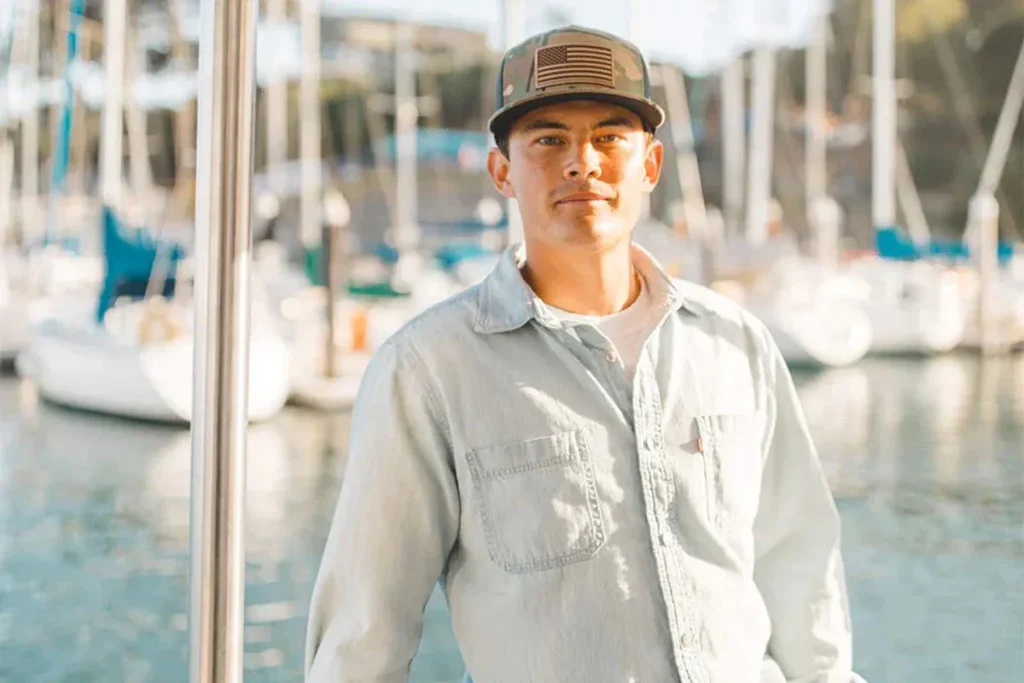 Man wearing camouflage trucker hat and light denim shirt by marina with boats in background.