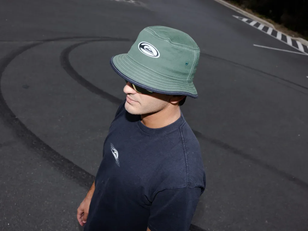 Man wearing green bucket hat and sunglasses standing on asphalt road.
