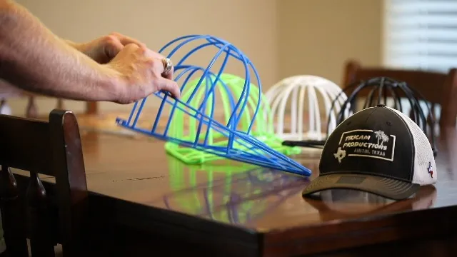 Hand holding blue hat shaping frame on wooden table with baseball cap nearby