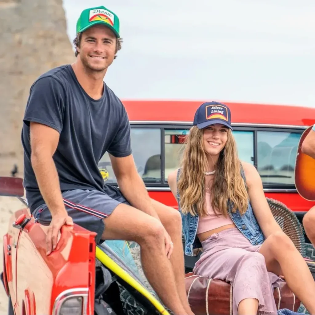 Young man and woman wearing colorful trucker hats sitting on red pickup truck.