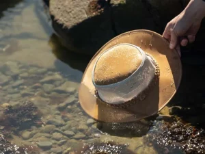 Hand holding wet brown cowboy hat partially submerged in clear water over stones.
