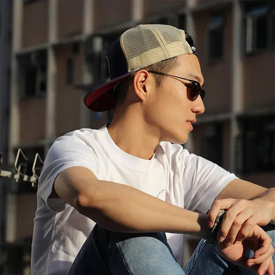 Young man wearing sunglasses and beige mesh trucker hat sitting outdoors.
