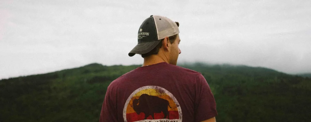 Man wearing beige and black trucker hat backward with nature view in background.