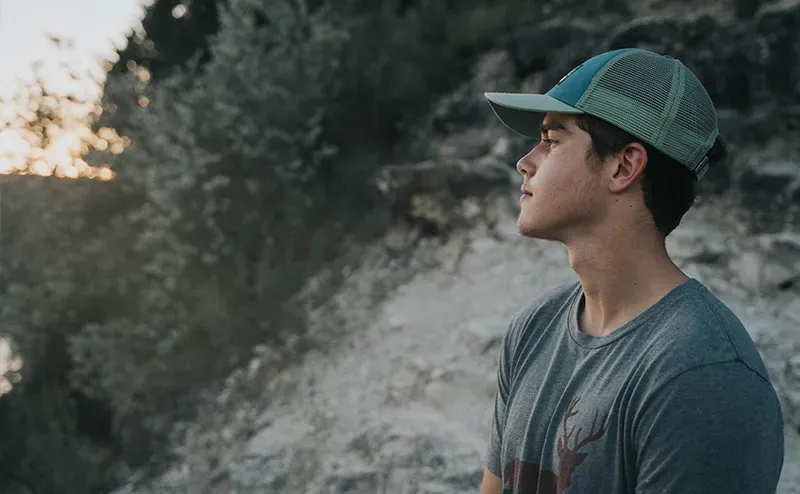 Young man in gray t-shirt wearing green trucker hat looking at rocky outdoors at sunset.