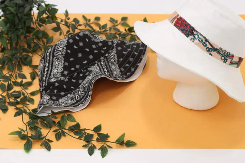 Two bucket hats, one black with white paisley print, one white with colorful patterned band, displayed with green leaves.