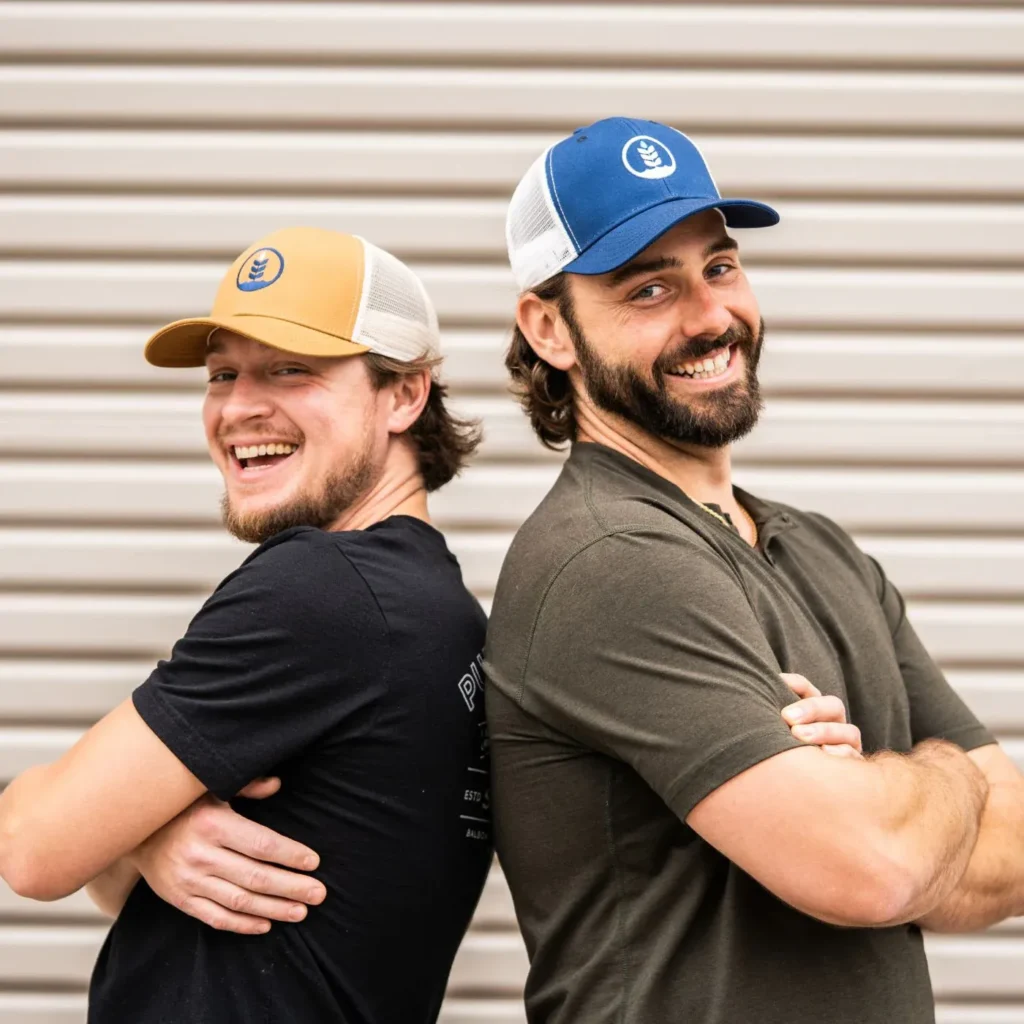 Two smiling men standing back to back wearing blue and yellow trucker hats.
