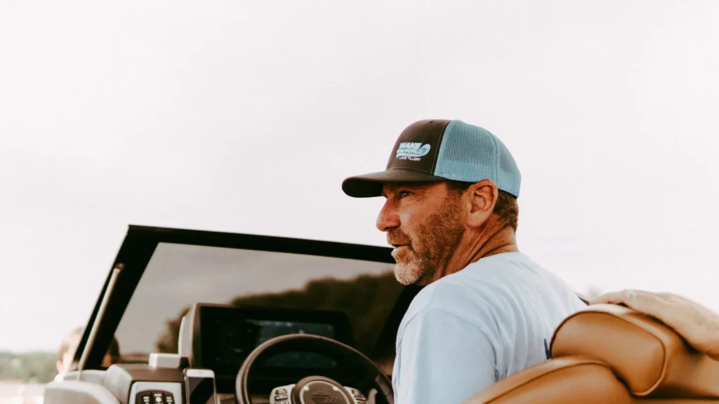 Middle-aged man driving boat wearing blue and brown trucker hat outdoors.