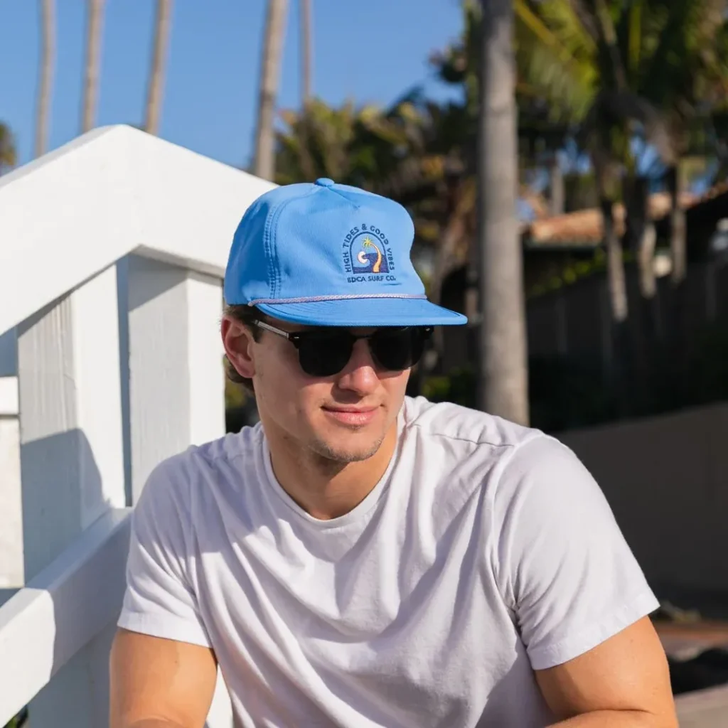 Man wearing sunglasses and blue baseball hat sitting outdoors near white railing