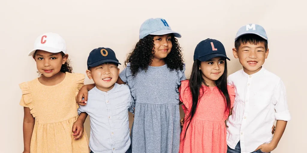 Group of five smiling children wearing colorful baseball caps with letters.