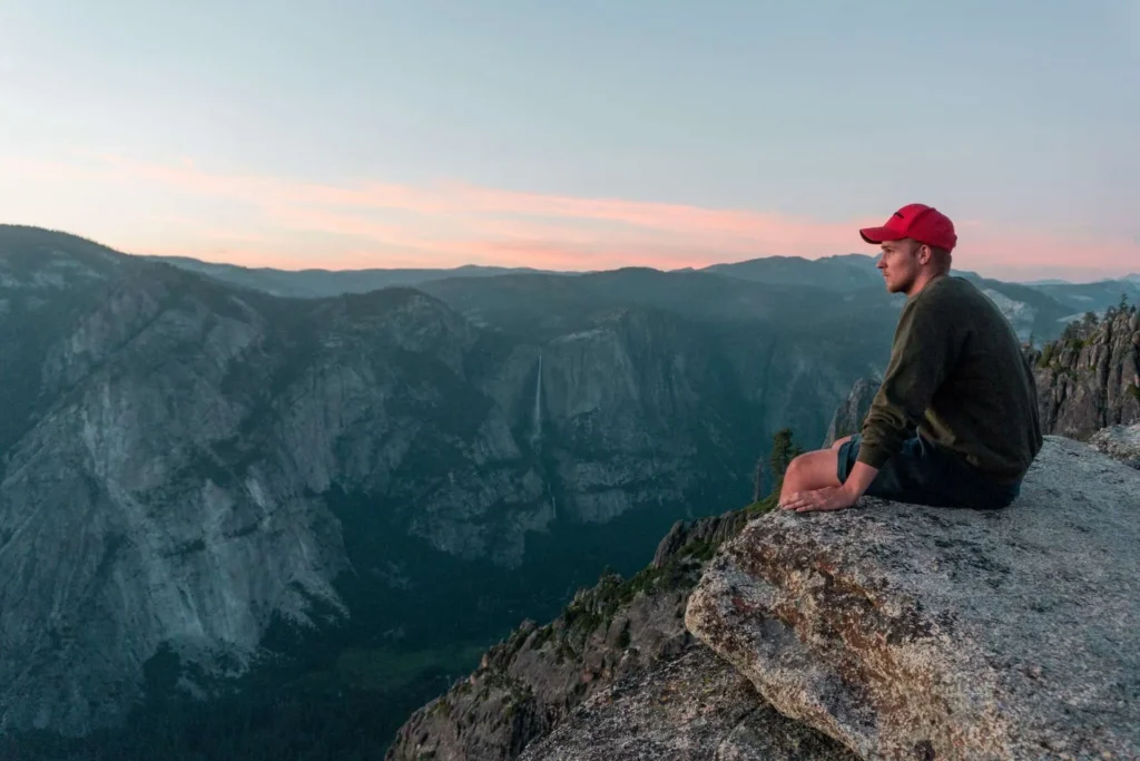 Man in red cap sitting on cliff edge overlooking mountains
