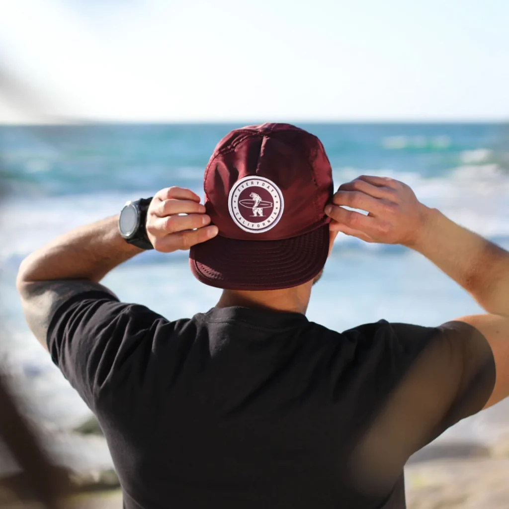 Man adjusting maroon cap with surf logo near ocean shore