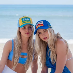 Two blonde women wearing colorful trucker hats posing on a sunny beach with ocean background.