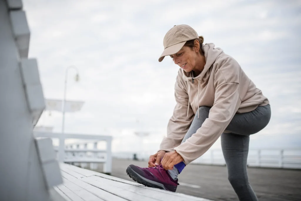 A woman outdoors in a beige cap, hoodie, and leggings, tying her running shoes with a joyful expression.