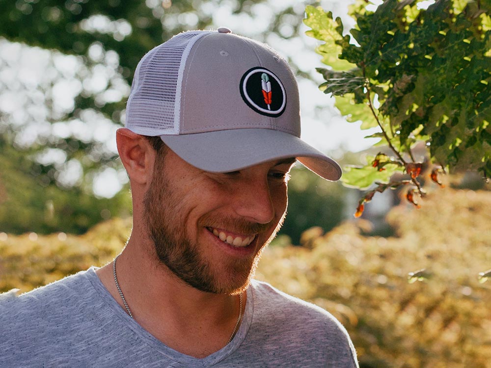 Smiling man wearing gray trucker hat with white mesh back outdoors.